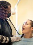 In bright room painted tan, a young girl with black pigtails smiles and looks up at a nurse practitioner as the NP holds a stethoscope to her back at Woods Healthcare's Medical Center.